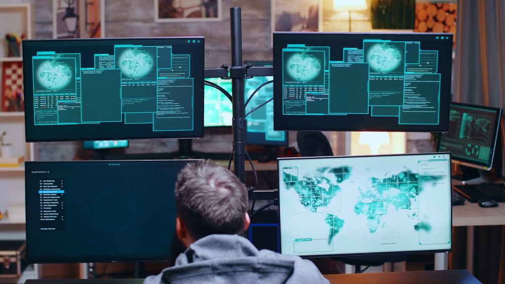 a picture of a hacker behind a desk with several computer screens – he is performing a cyber attack.
